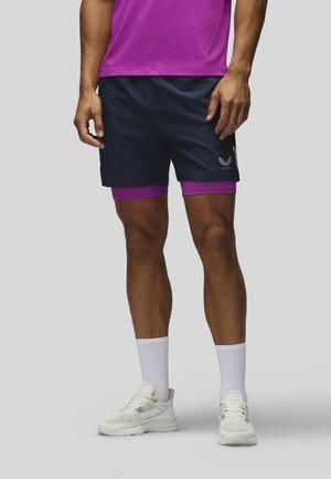 Man draagt zwarte sportshorts over paarse compressieshorts, witte sokken en witte hardloopschoenen, staat op een grijze achtergrond.