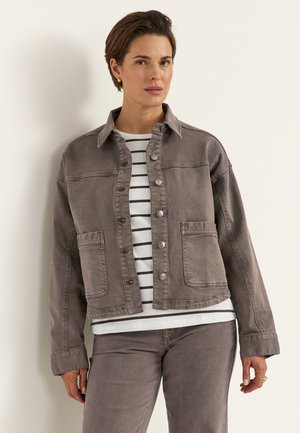 Kvinde iført en grå knappet jakke over en hvid og sort stribet skjorte, kombineret med matchende grå bukser, stående mod en beige væg.