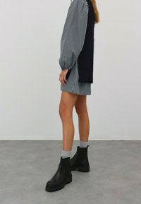 Schwarze elastische Chelsea Boots mit dicken, strukturierten Sohlen. Das Model trägt ein kariertes Kleid mit Puffärmeln und einem dunklen Strick-Back, kombiniert mit grauen Socken.