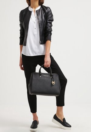 Femme portant une veste en cuir noire, un t-shirt henley blanc, un pantalon noir, des chaussures noires sans lacets, tenant un sac à main Michael Kors noir par les poignées.