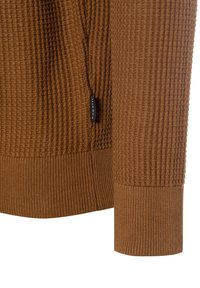 Brauner Strickpullover mit strukturiertem Rippenmuster und einem glatten, breiten Bündchen. Verfügt über einen seitlichen Schlitz und ein kleines Markenetikett an der Naht.