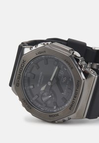 Montre G-SHOCK avec un bracelet en caoutchouc noir, un boîtier octogonal en acier inoxydable, un cadran sombre, des aiguilles argentées et un affichage numérique. Les caractéristiques incluent des boutons pour l'éclairage et le mode.