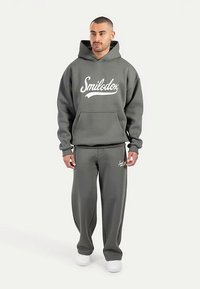 Sudadera con capucha gris y pantalones a juego hechos de tela suave. La sudadera presenta un bolsillo frontal y un gran logo blanco de "Smilodox". Las zapatillas blancas completan el conjunto.
