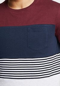 Muž v tričku s barevným blokováním v burgundské, námořnické modré, černé a bílé pruhy, které má na hrudi námořnickomodrou kapsu.