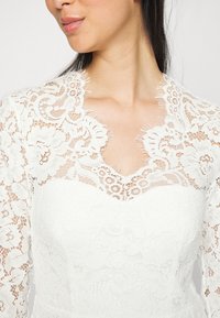 Valkoinen pitsimekko, jossa on V-muotoinen pääntie, jonka reunat ovat risaiset, monimutkaisia kukkakuoseja ja pitkät hihat. Sileä pinta koko mekossa.