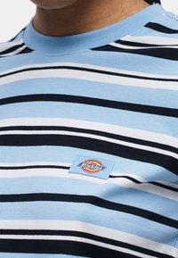 Κοντινό πλάνο ενός ατόμου που φοράει ένα ριγέ μπλουζάκι σε ανοιχτό μπλε, άσπρο και μαύρο με ένα μικρό ορθογώνιο μπατζάκι με το λογότυπο Dickies στο στήθος.