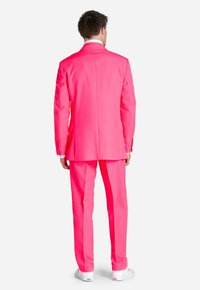 Completo rosa brillante con vestibilità su misura, caratterizzato da una fessura singola sul retro. Indossato con sneakers bianche per un contrasto informale.