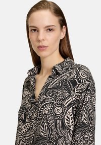 Langarmshirt van zwarte stof met een beige botanisch patroon, kraag en knoopsluiting, met een ontspannen pasvorm en zachte textuur.