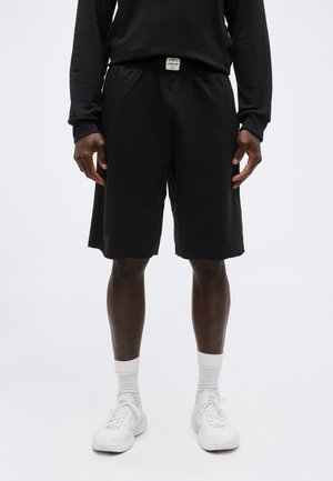 Άτομο που φοράει μαύρο σορτς μέχρι το γόνατο, μαύρο πουκάμισο με μακριά μανίκια, λευκές κάλτσες και λευκά αθλητικά παπούτσια, στέκεται μπροστά σε απλό λευκό φόντο.