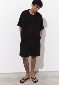Jeune homme aux cheveux bouclés se tenant debout et regardant vers le bas, portant une chemise noire à manches courtes boutonnée, un short noir et des tongs noires.