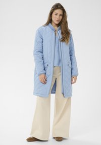 Svetlo modra prešita dolga jakna z zadrgo spredaj, dvema stranskima žepoma in rebrastim ovratnikom, kombinirana z bež širokimi hlačami in rjavimi čevlji.