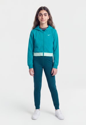 Fille debout de face portant un sweat à capuche zippé Nike turquoise, un pantalon assorti et des baskets blanches sur un fond clair uni.