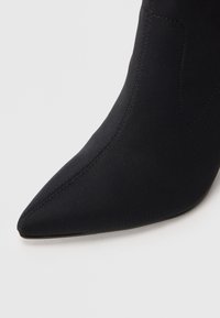 Czarne botki za kostkę z ostro zakończonym noskiem, bezszwowym designem oraz subtelnymi detalami szycia. Materiał jest gładki, co zapewnia elegancki wygląd.