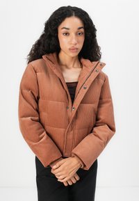 Woman with long curly hair wearing a brown corduroy puffer jacket over a dark top, standing against a plain white background.