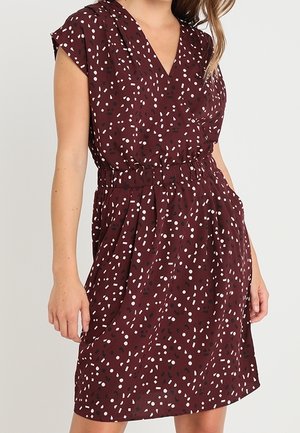 Nainen, jolla on ylipolvinen viininpunainen mekko, jossa on valkoista ja mustaa abstraktia konfettikuviota ja joustava vyötärö, seisoo rentoine käsineen.