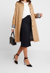 Trench-coat beige avec des fermetures à boutons et une doublure à carreaux sur les poignets, associé à une jupe ajustée foncée et des escarpins noirs à bride cheville. Sac à main noir inclus.
