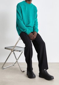 Turkos stickad tröja med ribbad manschett, i kombination med svarta vida byxor och svarta stövlar. Modell som sitter på en kromfällstol.