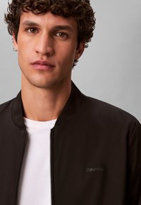 Veste bomber noire en tissu lisse, dotée d'une fermeture éclair et d'un col montant. Comprend un logo discret sur la poitrine, portée par-dessus une chemise blanche.