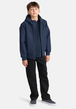 Marineblaue Kapuzenjacke mit Reißverschluss und gefüttertem Innenraum, kombiniert mit schwarzen Denim-Hosen und grauen Turnschuhen.