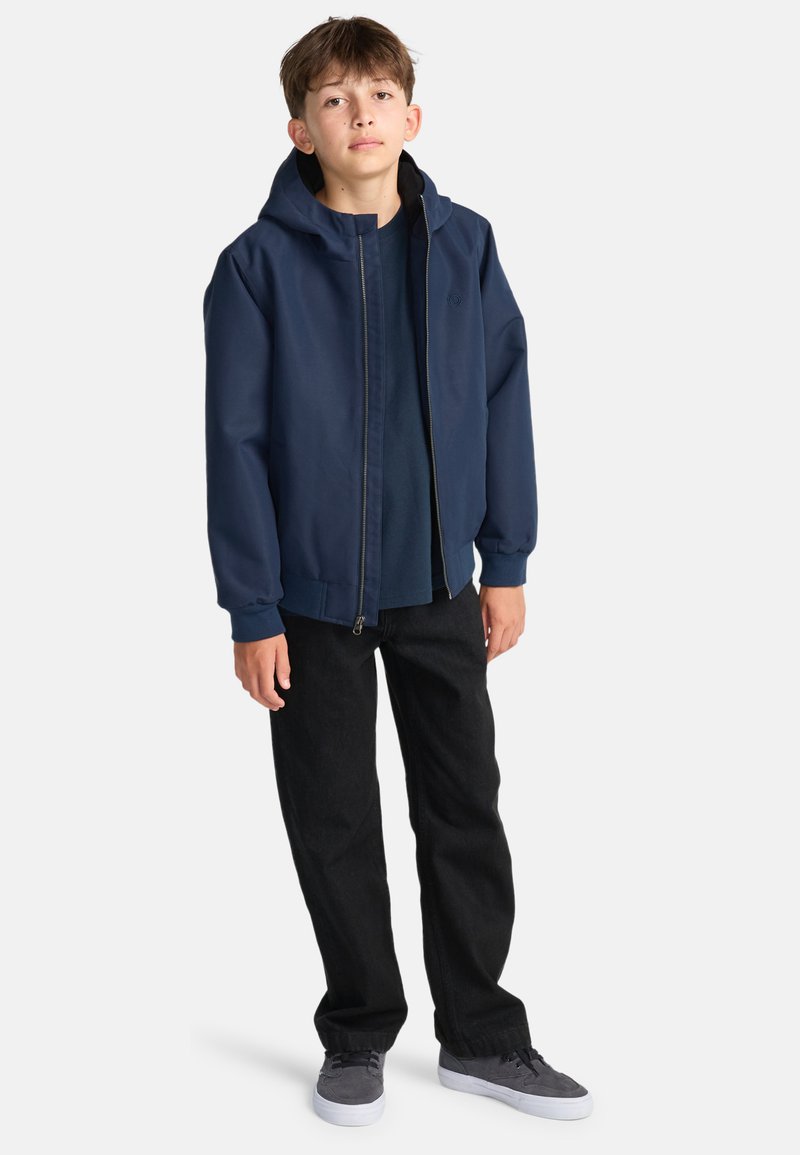 Giacca con cappuccio di colore blu navy, con zip frontale e una fodera interna, abbinata a pantaloni in denim neri e sneakers grigie.
