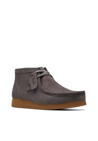 Bota de tobillo de ante gris con puntera redondeada, suela de goma marrón y costuras decorativas. Cuenta con un cierre de cordones y una etiqueta con el logotipo.