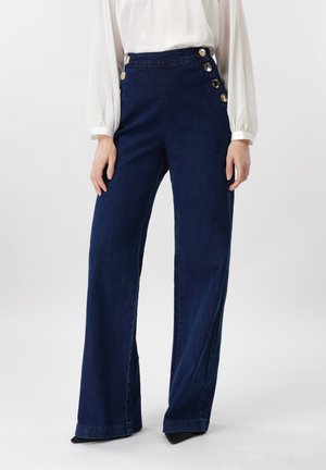 Γυναίκα που φορά ψηλόμεσο σκούρο μπλε παντελόνι φαρδιάς γραμμής με χρυσά κουμπιά στο πλάι και λευκό μακρυμάνικο πουκάμισο, στέκεται μπροστά σε λευκό φόντο.