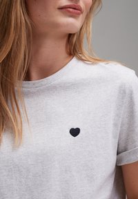 Graues Baumwoll-T-Shirt mit rundem Ausschnitt, kurzen umgeschlagenen Ärmeln und einer kleinen marineblauen Herzstickerei auf der linken Brust. Glatte Textur.