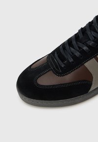 Black suede sneaker with brown leather accents, lace-up design, visible stitching, and curved, textured rubber sole. Subtle color blocking.