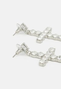 Boucles d'oreilles en croix couleur argent, ornées de strass transparents, avec un support et un clip robustes, affichant une texture lisse et brillante.