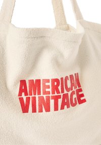 Sac fourre-tout couleur crème avec une surface douce et texturée. Présente une inscription en lettres rouges audacieuses "AMERICAN VINTAGE". Les poignées sont �épaisses et solides.