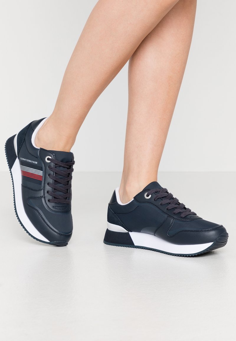 Nogi w czarno-białych sneakersach Tommy Hilfiger z czerwonymi i białymi paskami, stojące na jasnej podłodze na białym tle.