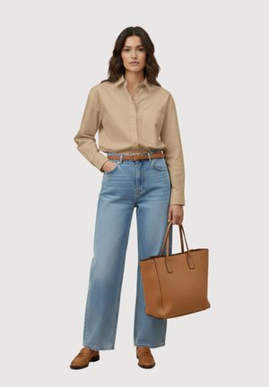 Vrouw die een beige shirt met lange mouwen draagt, blauwe high-waist jeans, een bruine riem, bruine loafers en een grote bruine handtas vasthoudt.