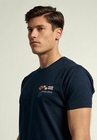 Camiseta de algodón azul marino con el texto "SAILING" y diseños de bandera en el pecho, con un clásico cuello redondo y mangas cortas.