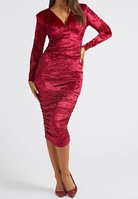 Robe moulante en velours rouge avec manches longues, texture froncée, décolleté en V profond, ourlet à hauteur des genoux, associée à des talons pointus rouges assortis.