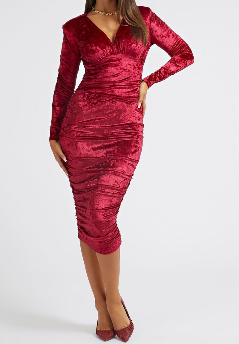 Robe moulante en velours rouge avec manches longues, texture froncée, décolleté en V profond, ourlet à hauteur des genoux, associée à des talons pointus rouges assortis.