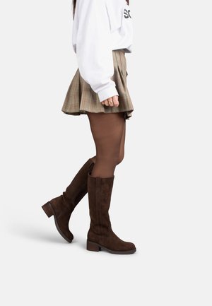 Vrouw draagt bruine suède kniehoge laarzen, doorschijnende donkere panty's en een geplooide geruite rok, staat met één been iets omhoog.