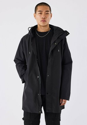 Manteau long noir imperméable avec une capuche, fermeture éclair et boutons-pression, coupe décontractée et poches latérales. Porté sur un haut noir.