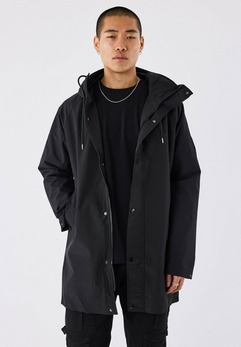 Giacca lunga nera impermeabile con cappuccio, chiusure a zip e a scatto, vestibilità comoda e tasche laterali. Indossata sopra un top nero.