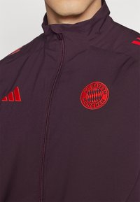 Bordeaux sportsjakke med høy krage, prydet med rødt brodert FC Bayern München-logo og røde Adidas-detaljer på skuldrene.