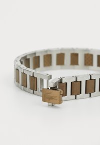 Bracelet en métal composé de maillons en argent et d'accents en bronze rectangulaires. Dispose d'un fermoir avec un logo gravé. Texture lisse et design élégant.