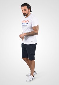 T-shirt en coton blanc avec du texte bleu et rouge, associé à un short cargo marine. Des baskets complètent le look décontracté. Un tatouage est visible sur le bras.