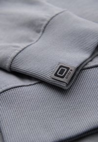 Gros plan d’un poignet de sweat en tissu côtelé gris avec une petite étiquette de marque tissée portant la mention « Ombre » cousue près de la couture.
