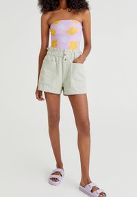 Zelené šortky s elastickým pasem a dvěma předními kapsami, doplněné fialovým topem bez ramínek s květinovým vzorem. Outfitu dominuje levandulové sandály.