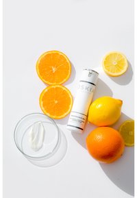 Il tubo di crema Oskia Renaissance 360 accanto a metà limone e arancia, con un piccolo piatto di vetro contenente una sostanza bianca cremosa.