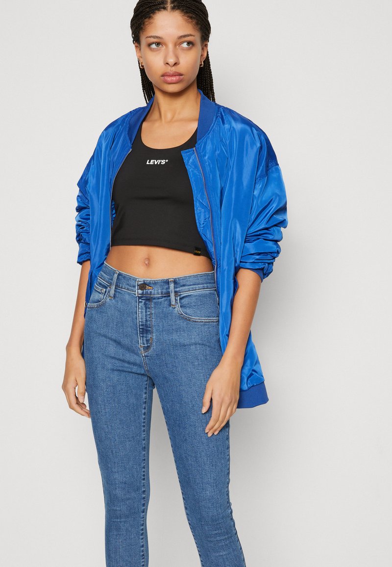 Blaues Bomberjackett mit satiniertem Finish, getragen über einem schwarzen Cropped-Top mit einem weißen Logo, kombiniert mit eng anliegenden blauen Jeans.