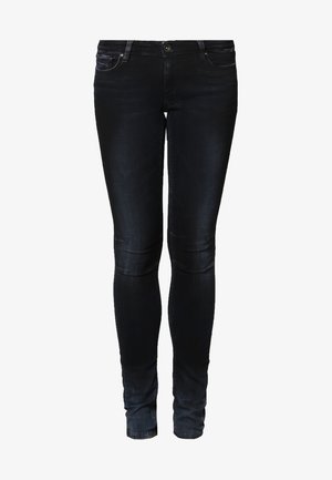 Pomarańczowa luźna koszulka z kieszenią, czarne dopasowane jeansy, białe buty slip-on oraz brązowy zamszowy plecak na sznurek z frędzlem.