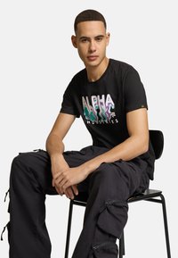 Musta puuvillainen t-paita, jossa on värikäs "ALPHA INDUSTRIES" -logo, yhdistetty mustiin cargo-housuihin, joissa on vetoketjuja ja taskuja, istuen mustassa tuolissa.