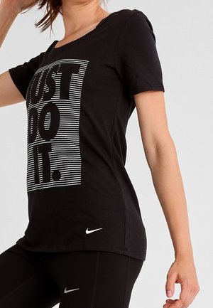 Kobieta w czarnej koszulce Nike z napisem "Just Do It." oraz czarnych legginsach z białymi logo Nike, pozująca z uniesionym ramieniem.