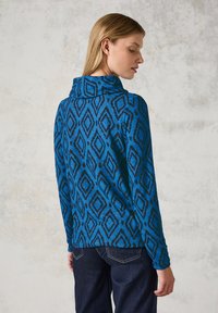 Blauwe gebreide trui met diamantvormen, hoge kraag en lange mouwen, gemaakt van een zacht materiaal, gedragen met donkere jeans.