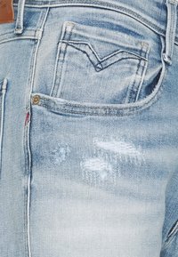 Světle modré džíny s zadní kapsou, která má tmavé švy a oprýskané okraje v oblasti kapsy a stehna.